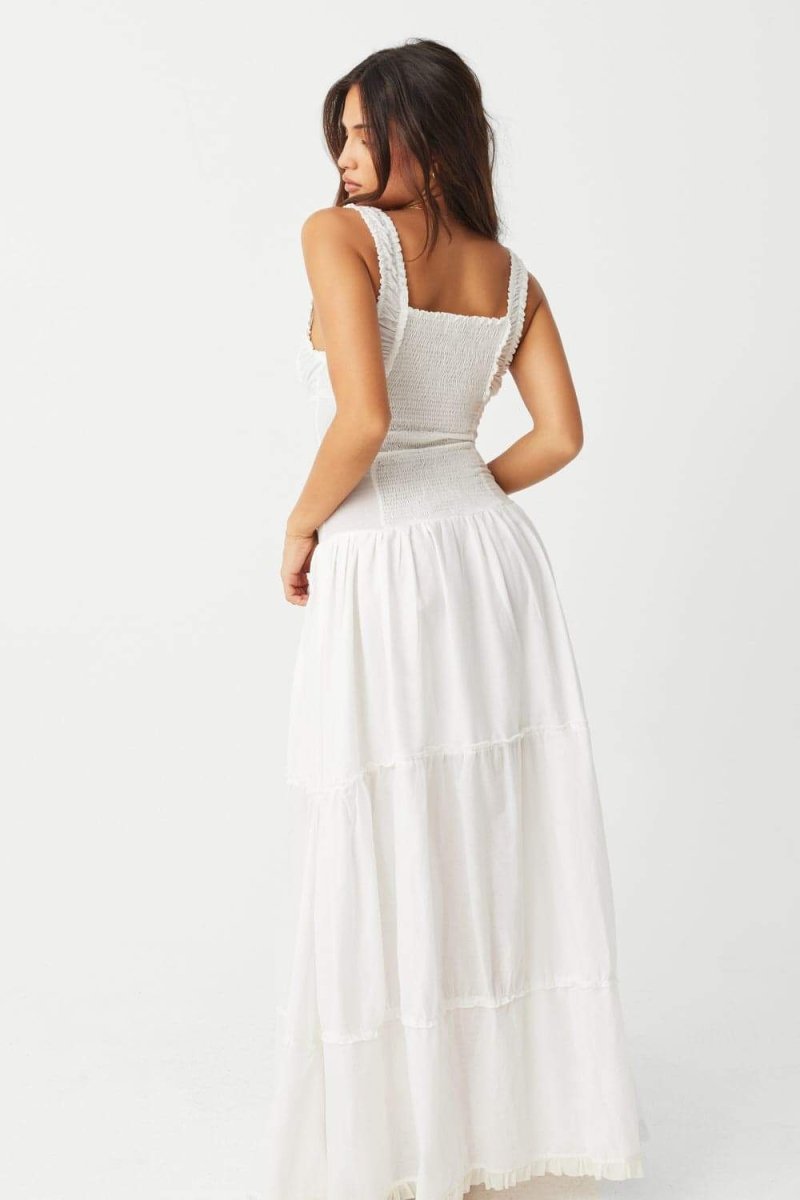 White - A woman sitting on rocks by the ocean wearing a white, sleeveless maxi dress featuring a ruched square neckline, corset-style buttoned bodice, and a voluminous tiered skirt.