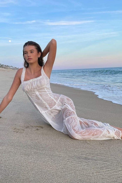 White - A woman sitting on rocks by the ocean wearing a white, sleeveless maxi dress featuring a ruched square neckline, corset-style buttoned bodice, and a voluminous tiered skirt.