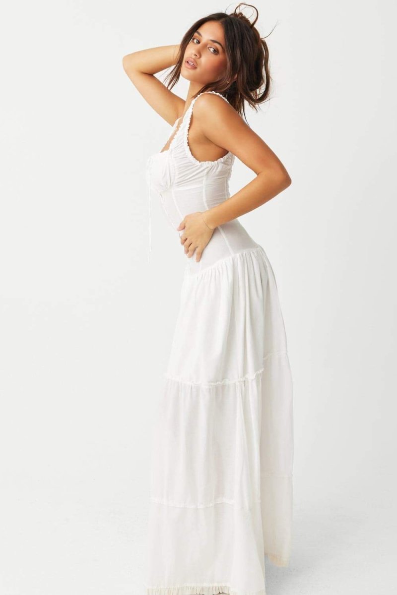 White - A woman sitting on rocks by the ocean wearing a white, sleeveless maxi dress featuring a ruched square neckline, corset-style buttoned bodice, and a voluminous tiered skirt.