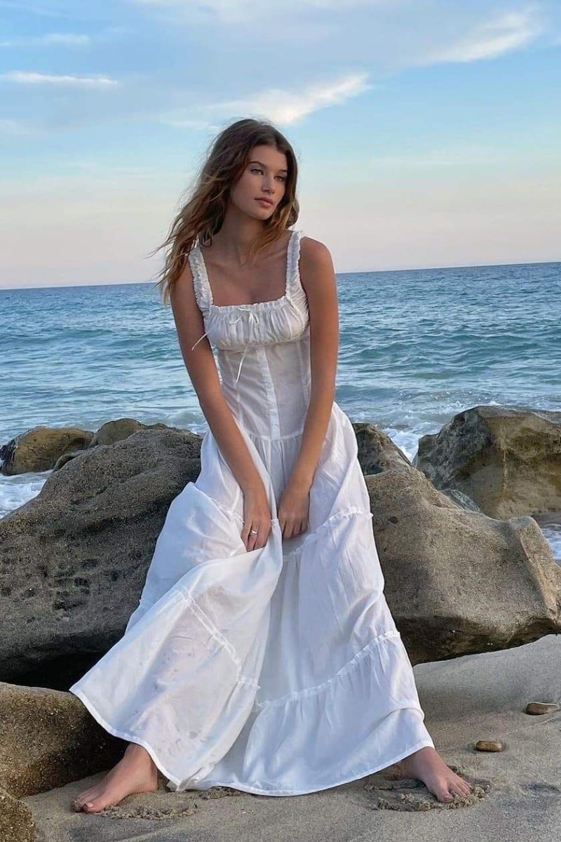 White - A woman sitting on rocks by the ocean wearing a white, sleeveless maxi dress featuring a ruched square neckline, corset-style buttoned bodice, and a voluminous tiered skirt.