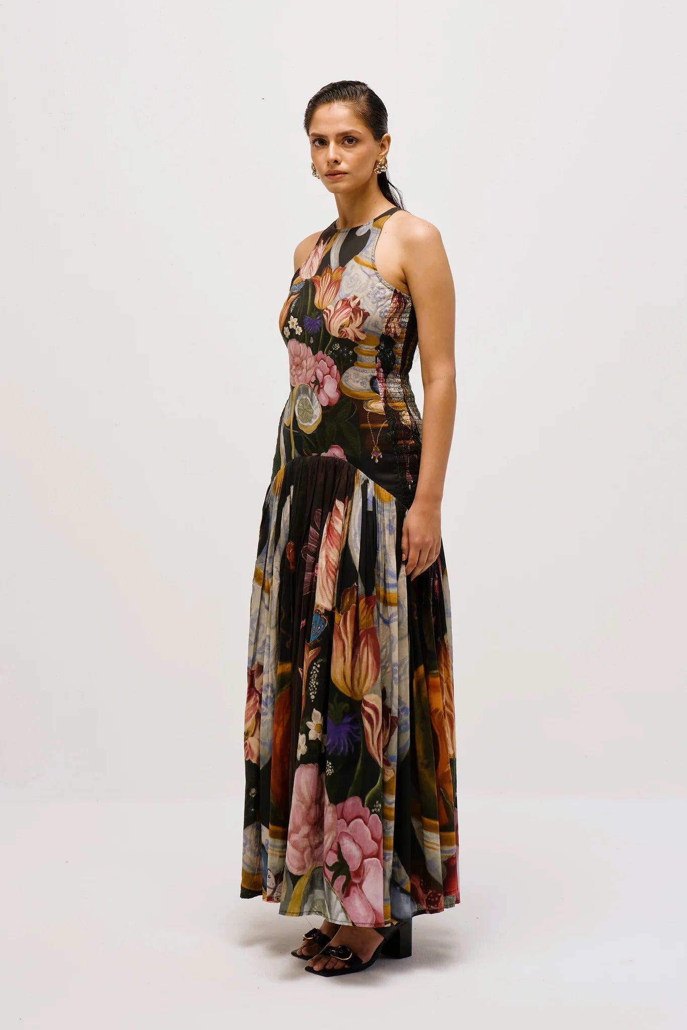 Multi - Full-length view of a woman wearing a sleeveless maxi dress with a high halter neckline and a pleated skirt, featuring a detailed still life floral and vase print on a dark background.
