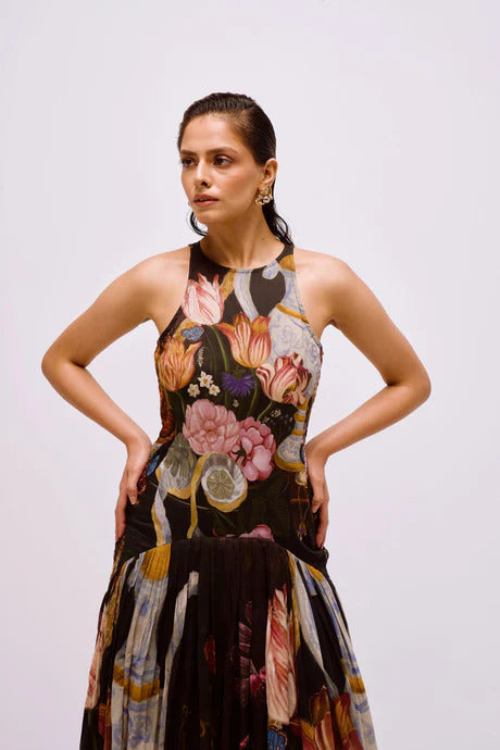 Multi - Full-length view of a woman wearing a sleeveless maxi dress with a high halter neckline and a pleated skirt, featuring a detailed still life floral and vase print on a dark background.