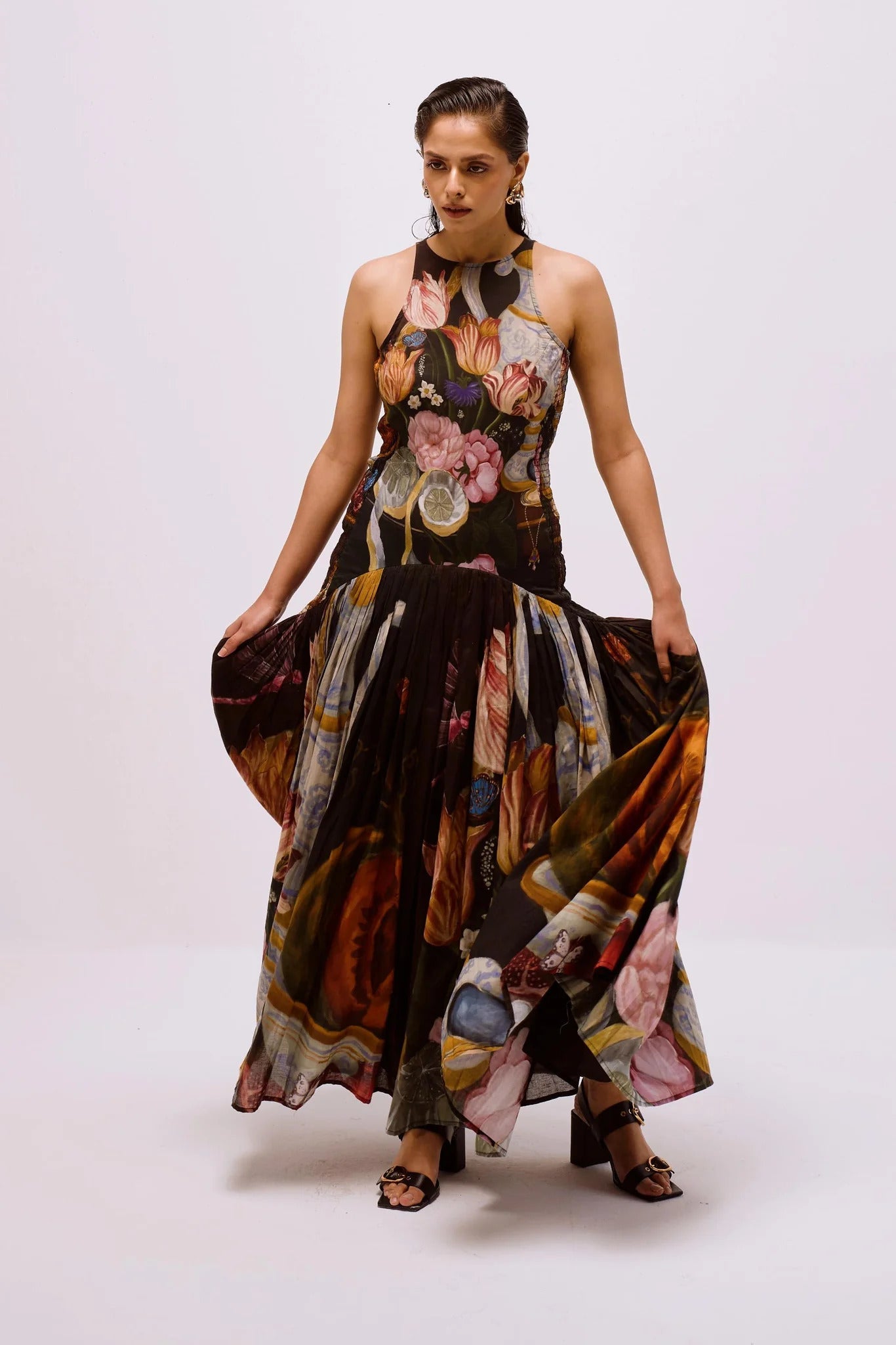 Multi - Full-length view of a woman wearing a sleeveless maxi dress with a high halter neckline and a pleated skirt, featuring a detailed still life floral and vase print on a dark background.