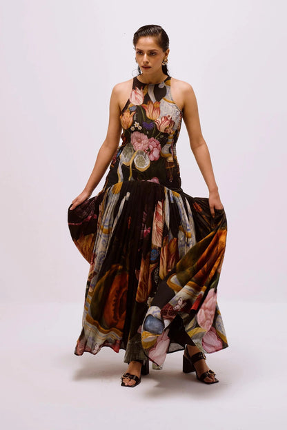Multi - Full-length view of a woman wearing a sleeveless maxi dress with a high halter neckline and a pleated skirt, featuring a detailed still life floral and vase print on a dark background.
