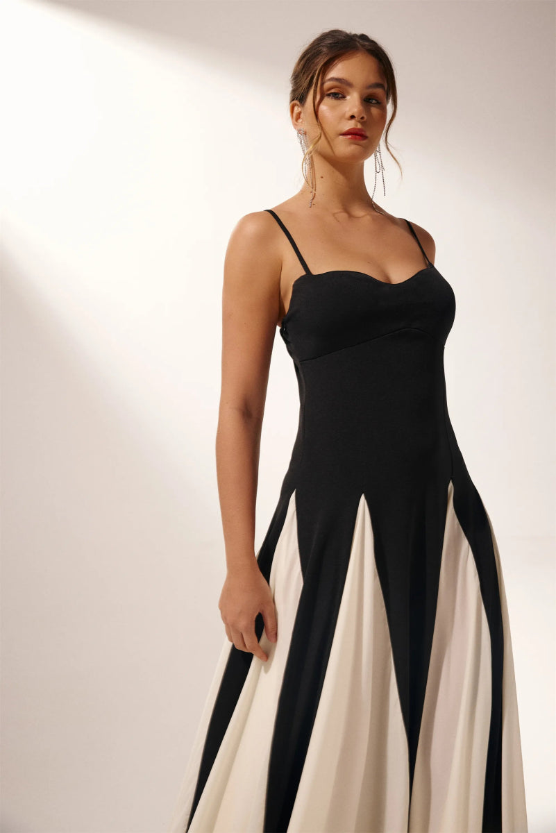 Black - A woman models a floor-length, two-tone maxi dress featuring a fitted dark bodice with spaghetti straps and a voluminous skirt constructed with alternating dark and flowing light godet panels.