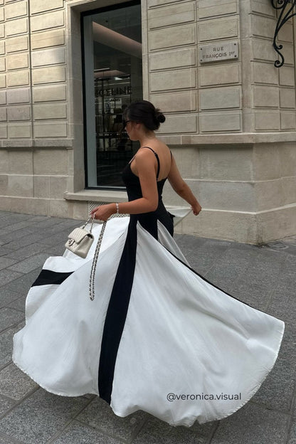Black - A woman models a floor-length, two-tone maxi dress featuring a fitted dark bodice with spaghetti straps and a voluminous skirt constructed with alternating dark and flowing light godet panels.