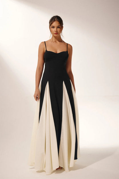 Black - A woman models a floor-length, two-tone maxi dress featuring a fitted dark bodice with spaghetti straps and a voluminous skirt constructed with alternating dark and flowing light godet panels.