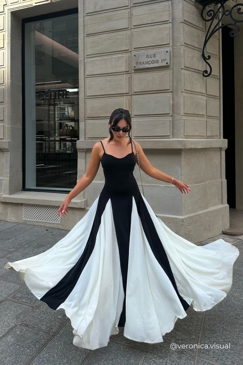 Black - A woman models a floor-length, two-tone maxi dress featuring a fitted dark bodice with spaghetti straps and a voluminous skirt constructed with alternating dark and flowing light godet panels.