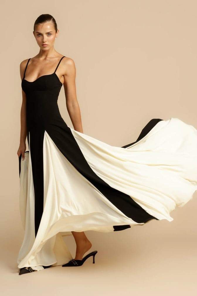 Black - A woman models a floor-length, two-tone maxi dress featuring a fitted dark bodice with spaghetti straps and a voluminous skirt constructed with alternating dark and flowing light godet panels.