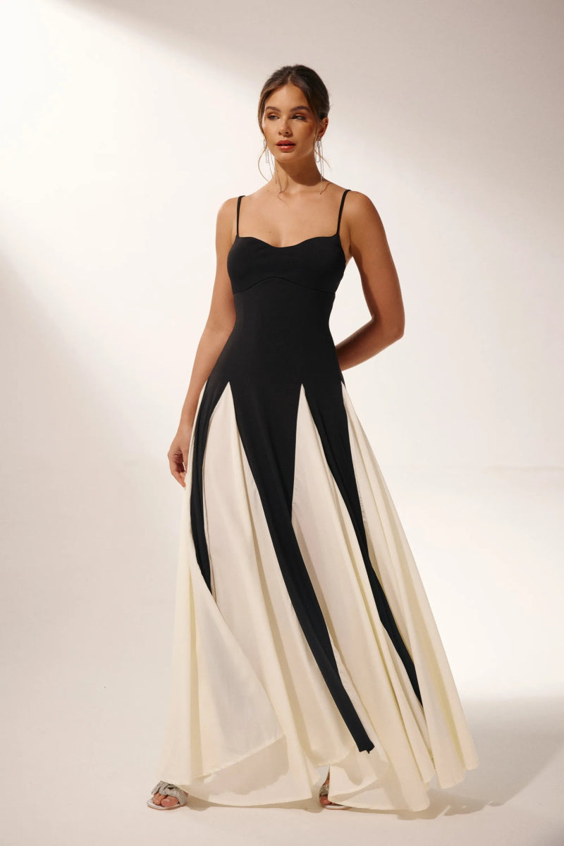 Black - A woman models a floor-length, two-tone maxi dress featuring a fitted dark bodice with spaghetti straps and a voluminous skirt constructed with alternating dark and flowing light godet panels.