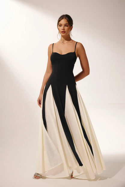 Black - A woman models a floor-length, two-tone maxi dress featuring a fitted dark bodice with spaghetti straps and a voluminous skirt constructed with alternating dark and flowing light godet panels.