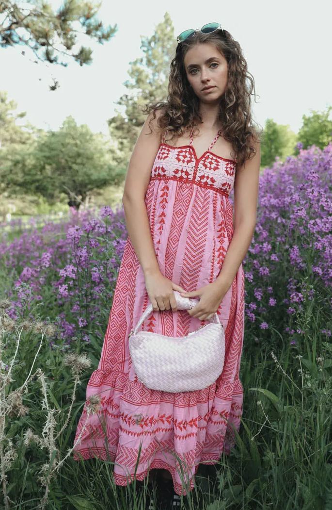 Woman standing in a field of purple flowers wearing a pink and red halter maxi dress with a geometric print, embroidered bodice, and tiered skirt.