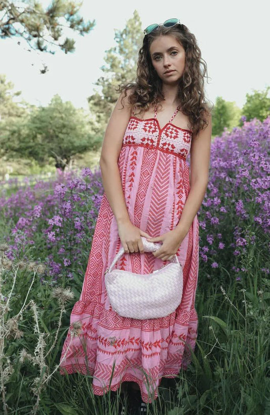 Woman standing in a field of purple flowers wearing a pink and red halter maxi dress with a geometric print, embroidered bodice, and tiered skirt.