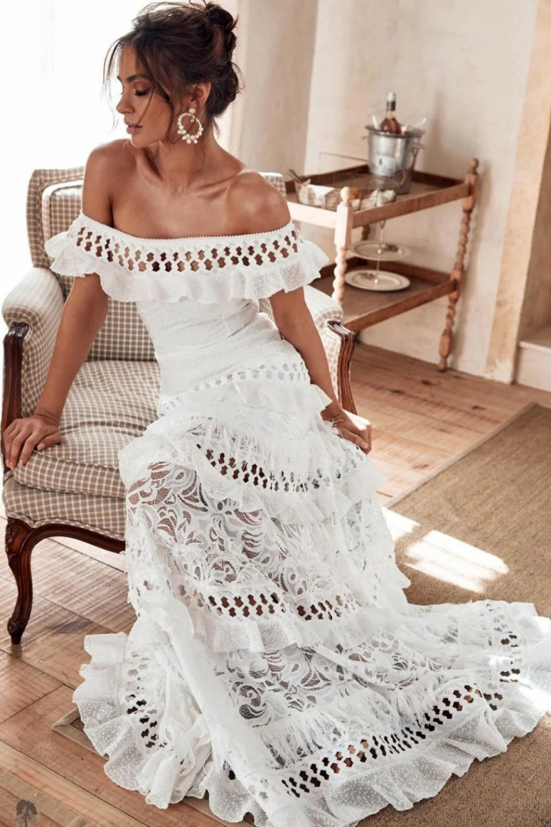 White - Woman wearing a white, off-the-shoulder maxi dress with tiered ruffles, lace, and broderie anglaise detailing.