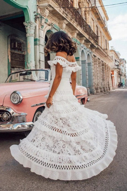 White - Woman wearing a white, off-the-shoulder maxi dress with tiered ruffles, lace, and broderie anglaise detailing.