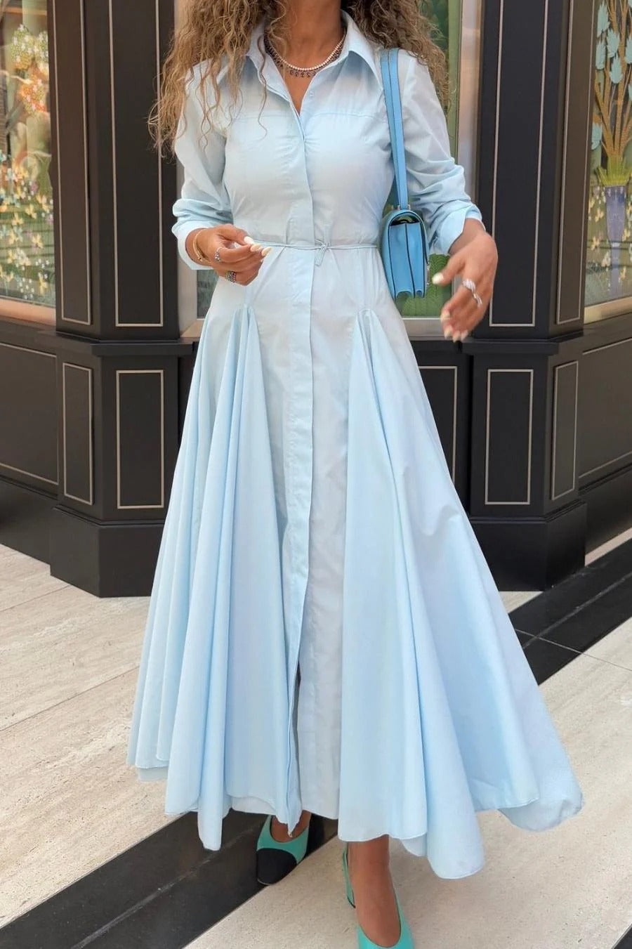 Lake Blue - A woman wearing a light blue, long-sleeved midi shirt dress featuring a collared neckline, button-down front, tie waist, and a voluminous pleated skirt with an asymmetric hem.
