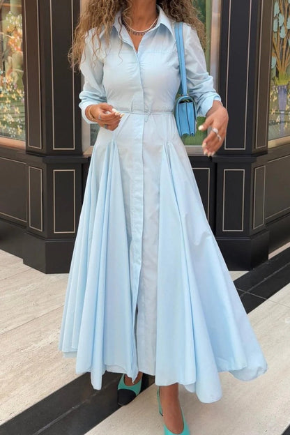 Lake Blue - A woman wearing a light blue, long-sleeved midi shirt dress featuring a collared neckline, button-down front, tie waist, and a voluminous pleated skirt with an asymmetric hem.