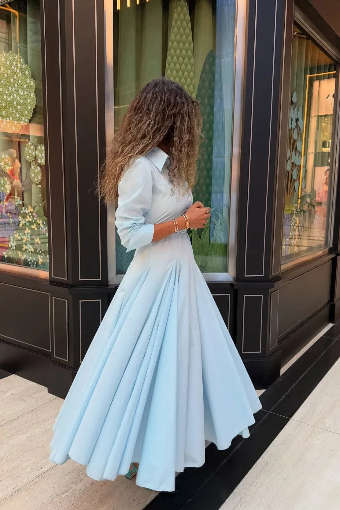 Lake Blue - A woman wearing a light blue, long-sleeved midi shirt dress featuring a collared neckline, button-down front, tie waist, and a voluminous pleated skirt with an asymmetric hem.
