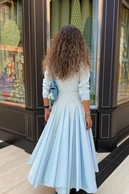 Lake Blue - A woman wearing a light blue, long-sleeved midi shirt dress featuring a collared neckline, button-down front, tie waist, and a voluminous pleated skirt with an asymmetric hem.