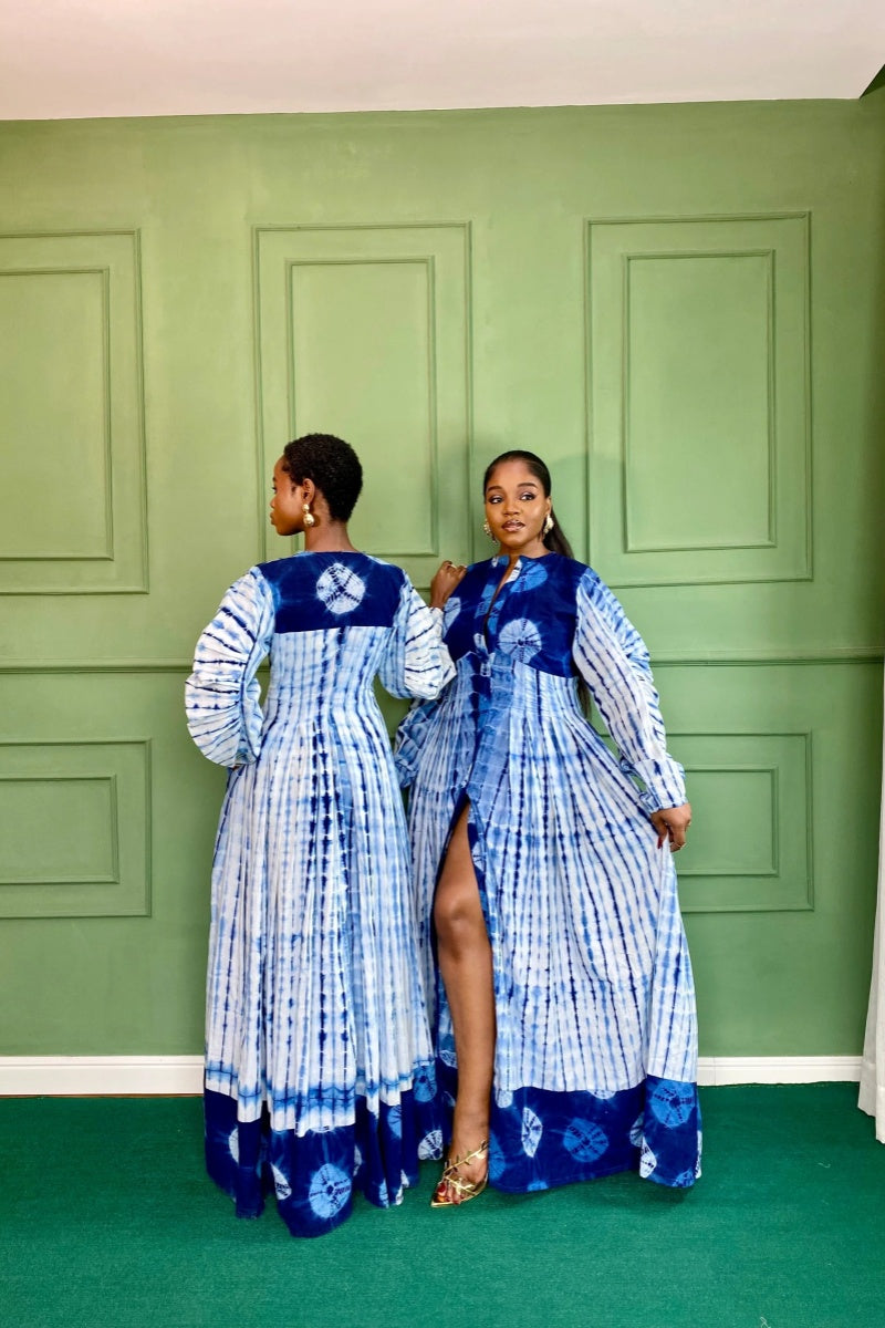 Blue - A woman wearing a long, blue and white shibori-style tie-dye maxi dress with long voluminous sleeves, a button-front closure, and a front slit, styled with white strappy heels.