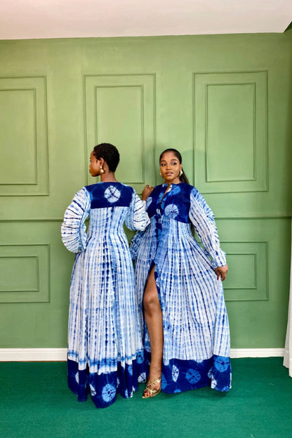 Blue - A woman wearing a long, blue and white shibori-style tie-dye maxi dress with long voluminous sleeves, a button-front closure, and a front slit, styled with white strappy heels.