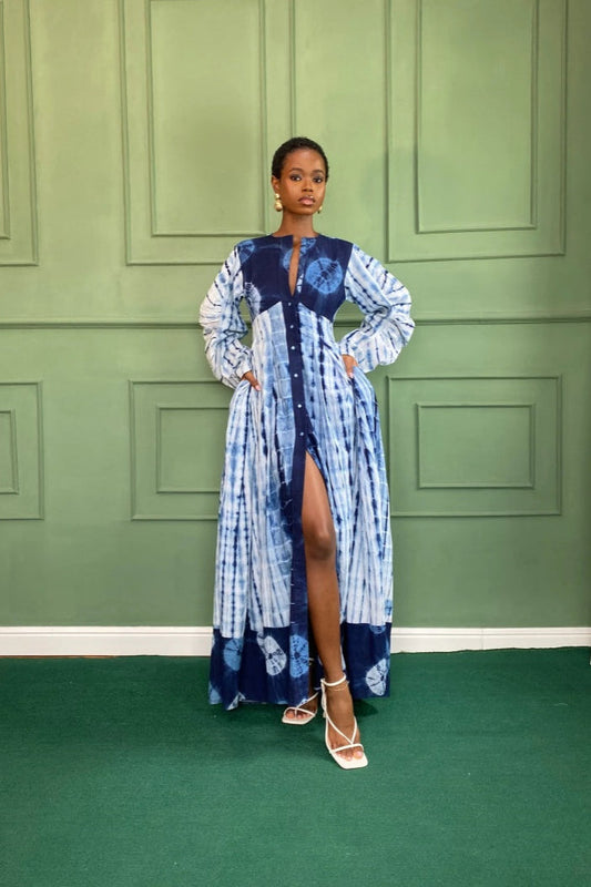 Blue - A woman wearing a long, blue and white shibori-style tie-dye maxi dress with long voluminous sleeves, a button-front closure, and a front slit, styled with white strappy heels.