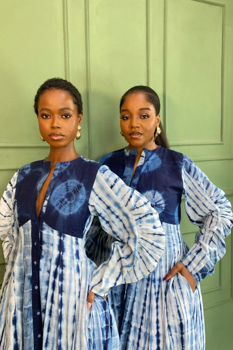 Blue - A woman wearing a long, blue and white shibori-style tie-dye maxi dress with long voluminous sleeves, a button-front closure, and a front slit, styled with white strappy heels.