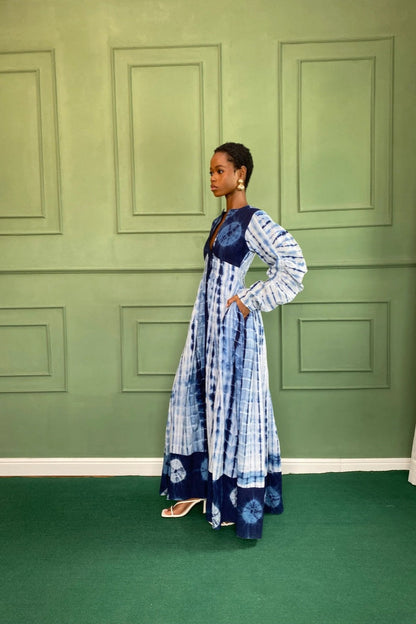 Blue - A woman wearing a long, blue and white shibori-style tie-dye maxi dress with long voluminous sleeves, a button-front closure, and a front slit, styled with white strappy heels.