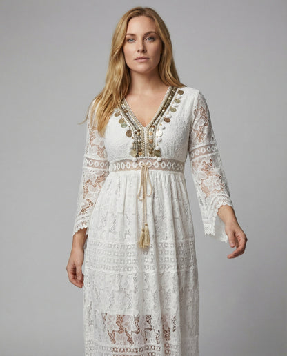 White - A woman wearing a bohemian lace maxi dress with a deep V-neckline, metallic embellishments, bell sleeves, and a tassel tie waist, holding a round woven bag.