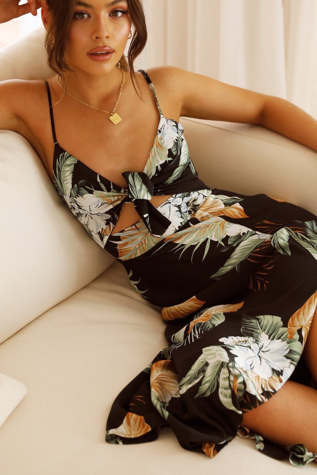 Black - A woman wearing a black midi dress with a bold tropical leaf and floral print, featuring a tie-front bodice, under-bust cut-out, spaghetti straps, and a flowing skirt with a front slit.
