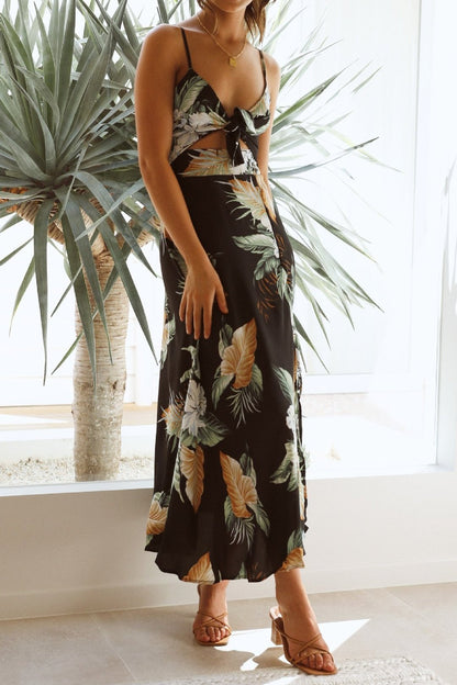 Black - A woman wearing a black midi dress with a bold tropical leaf and floral print, featuring a tie-front bodice, under-bust cut-out, spaghetti straps, and a flowing skirt with a front slit.
