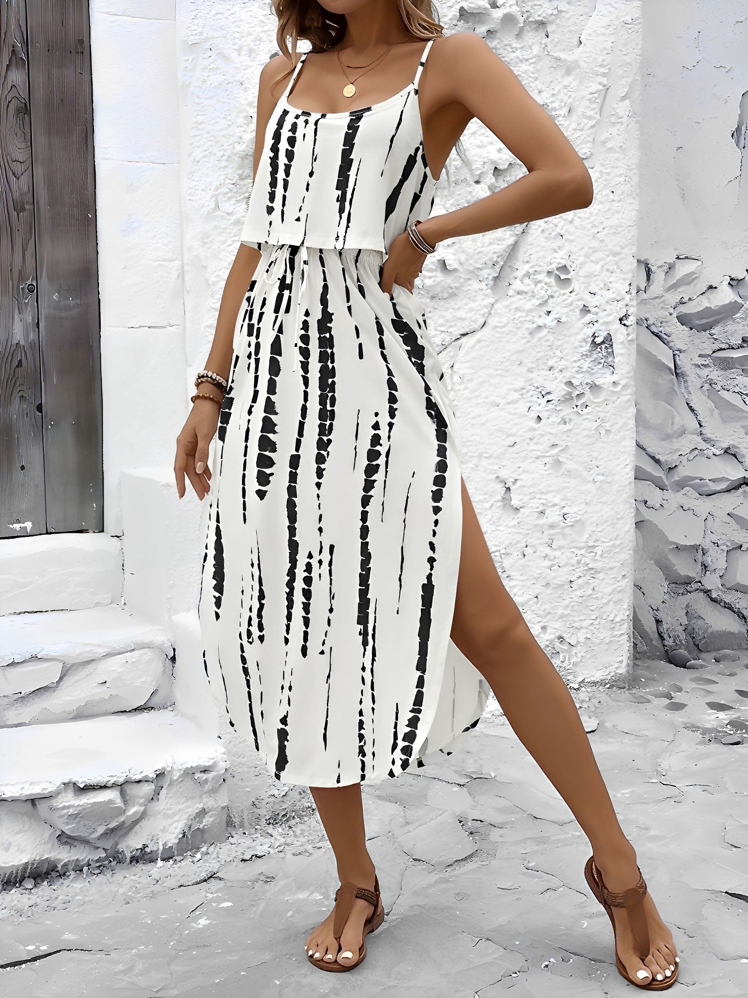 White - Model wearing a lightweight woven strappy midi dress with an abstract vertical pattern, adjustable waist tie, and side split, standing outdoors against a white textured wall.