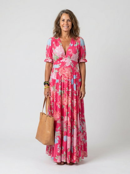Pink - A woman modelling a floral V-neck maxi dress with short sleeves and a tiered skirt, holding a structured tote bag against a light grey studio background.