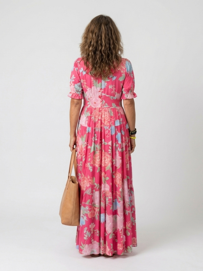 Pink - A woman modelling a floral V-neck maxi dress with short sleeves and a tiered skirt, holding a structured tote bag against a light grey studio background.