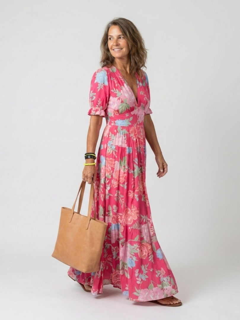 Pink - A woman modelling a floral V-neck maxi dress with short sleeves and a tiered skirt, holding a structured tote bag against a light grey studio background.