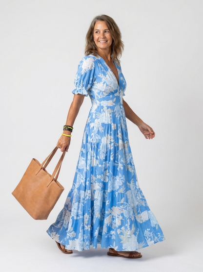 Light blue - A woman modelling a floral V-neck maxi dress with short sleeves and a tiered skirt, holding a structured tote bag against a light grey studio background.