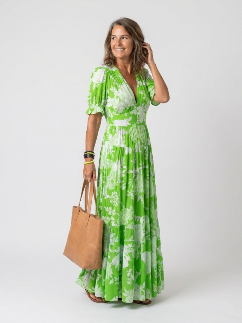 Green - A woman modelling a floral V-neck maxi dress with short sleeves and a tiered skirt, holding a structured tote bag against a light grey studio background.
