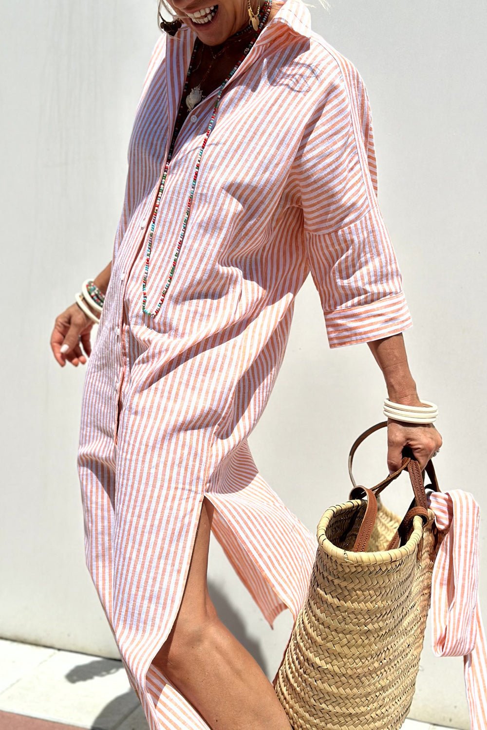 Pink - A woman wearing a striped, button-down midi-length shirt dress with 3/4 sleeves, carrying a woven straw basket bag outdoors.