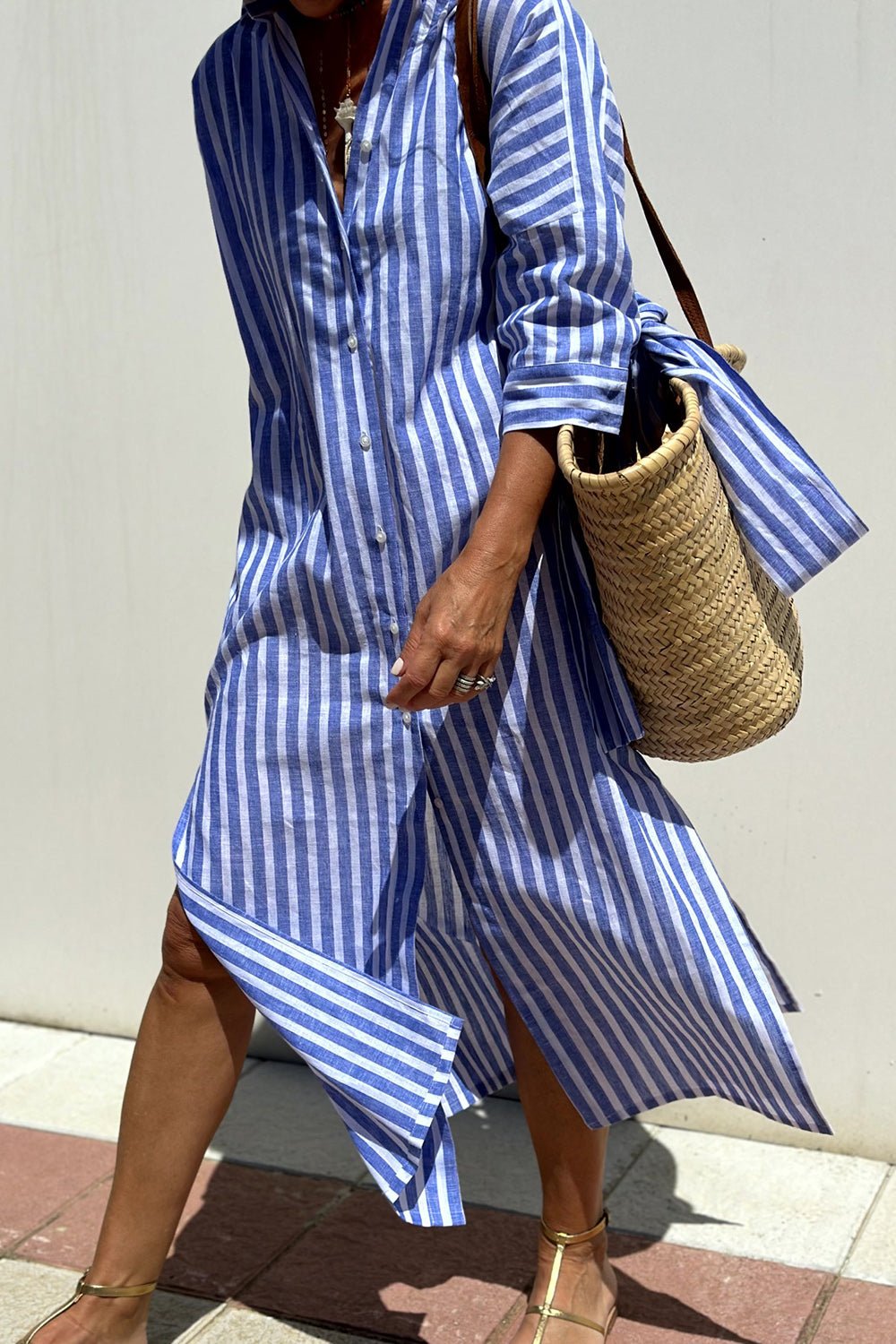 Blue - A woman wearing a striped, button-down midi-length shirt dress with 3/4 sleeves, carrying a woven straw basket bag outdoors.